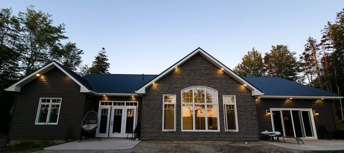 A newly built home in Southwest Nova Scotia at dusk, soffit lighting burning along every eave and the sunset caught in the arched front window.